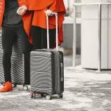 Dwie osoby w pomarańczowych kurtkach stoją z walizkami na zimnym, zaśnieżonym chodniku.